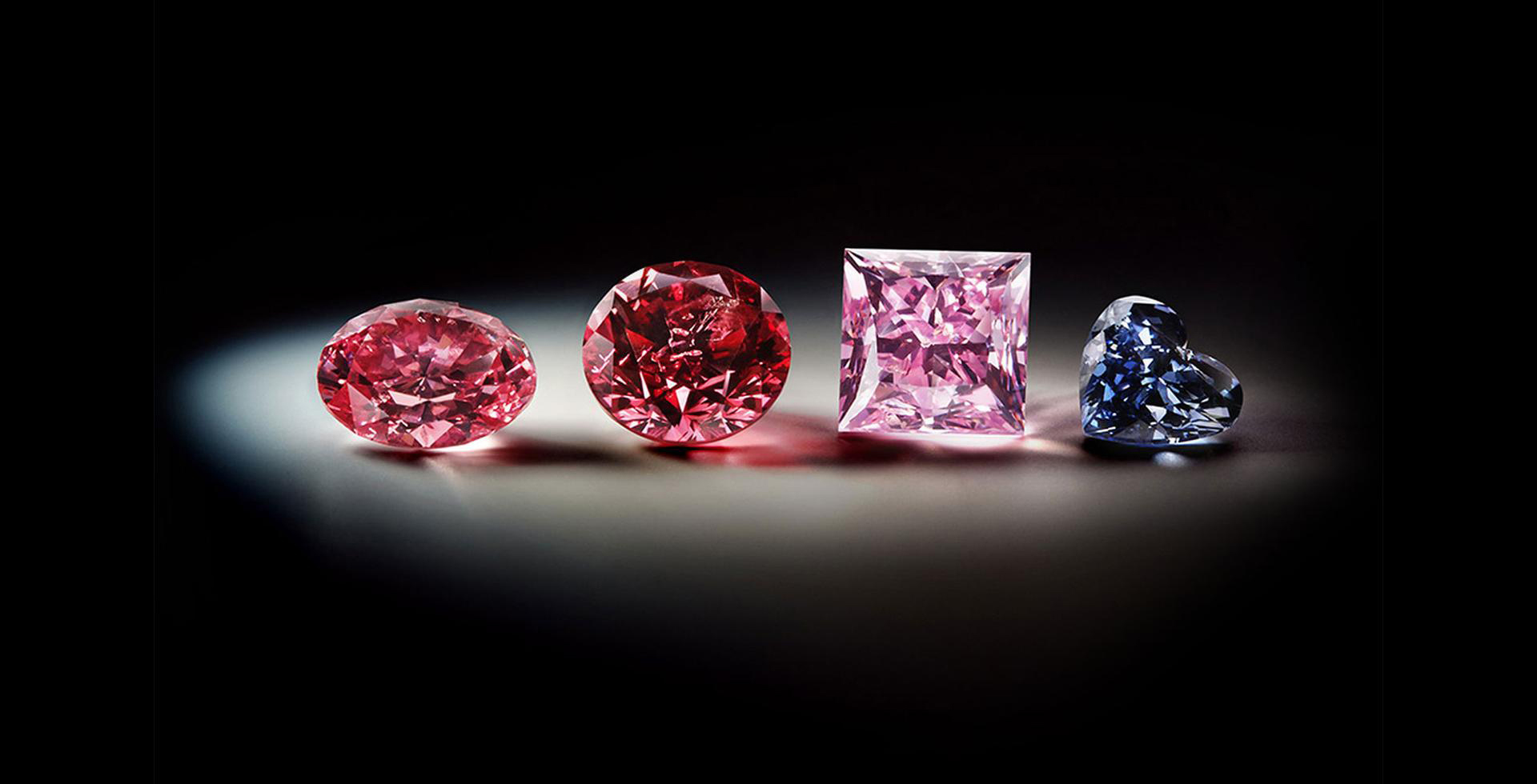 Four loose diamonds, two red, one pink and one blue heart shape, lit in a pool of light on a black ground