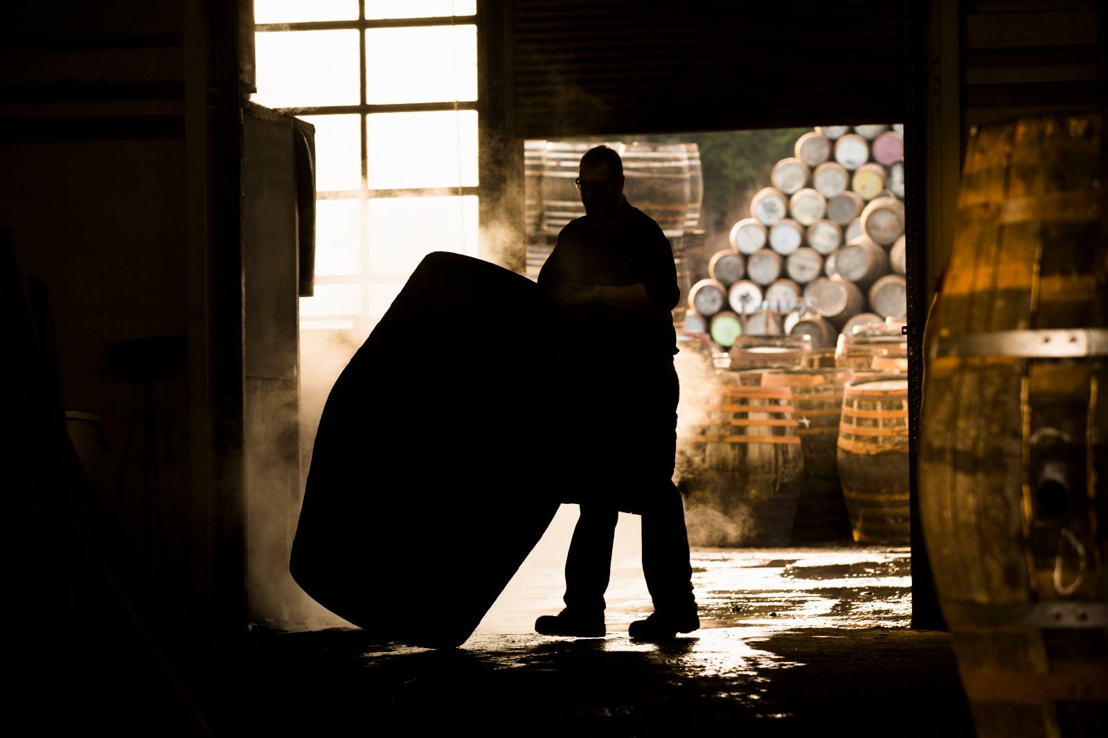 A silhouetted warehouse worker handling a cask in a doorway, stacked casks behind